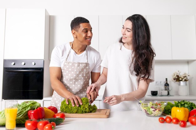 Foto coppia multirazziale prepara insalata vegetariana in cucina bianca ragazzo afroamericano in grembiule taglia i verdi