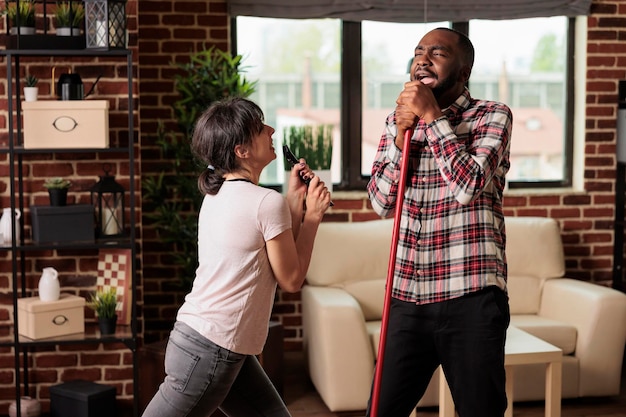 Multiracial couple singing at home using broom and tv remote control as microphone after finishing spring cleaning. Woman and African American man having fun with household chores.