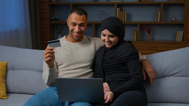 Multiracial couple sit on sofa use laptop purchasing on internet with credit card muslim wife woman