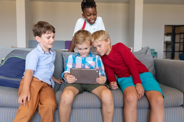 Photo multiracial elementary schoolboys looking at digital tablet while sitting on couch in school