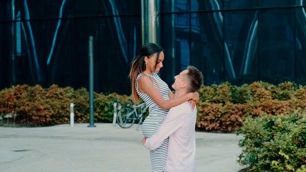 Multiracial family concept: husband spinning around his wife while walking down the street near big office centre one summer day