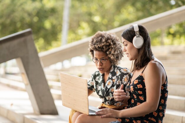 Multiracial female friends using laptop smiling looking at screen, wearing headphones and casual clothes, outdoors