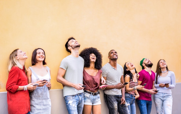 Photo multiracial friends having fun using smartphone at wall on university college break - young people addicted by mobile smart phones - technology concept with always connected millennials