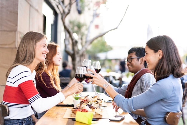 Multiracial friends toasting red wine at the outdoor pub in the\
city food and beverage lifestyle concept with happy people having\
fun together at open air pub bright filter with focus on\
glasses