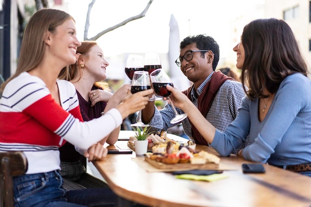 Multiracial friends toasting red wine at the outdoor pub food\
and beverage lifestyle concept with happy people having fun\
together at pub bright filter with focus on the guys in the\
background