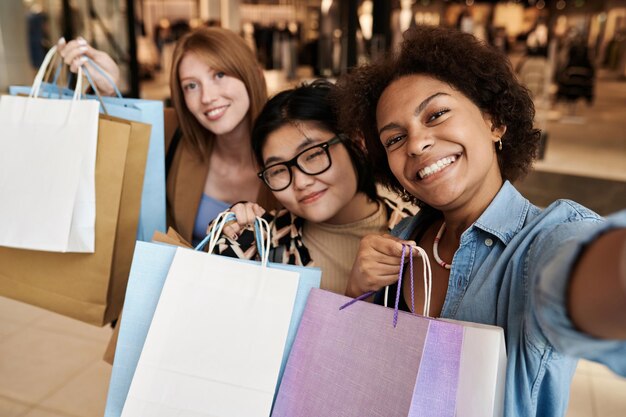 Photo multiracial group of friends taking selfie smiling at camera showing bags with purchases