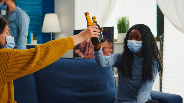 Multiracial group of friends with masks enjoying a beer during new normal party in living room respecting social distance to prevent virus spread. Diverse people enjoying free time in global pandemic