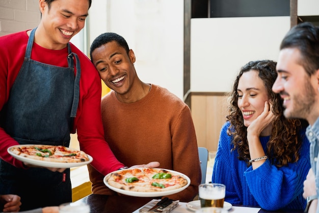エプロンを持ったアジア人の笑顔の男性が彼らにもたらしているピザと友達の多民族グループ