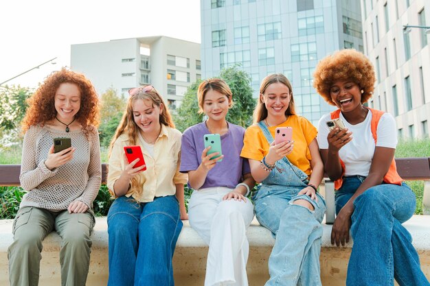 Photo multiracial group of young female friends enjoying and smiling using their mobile phone app sitting