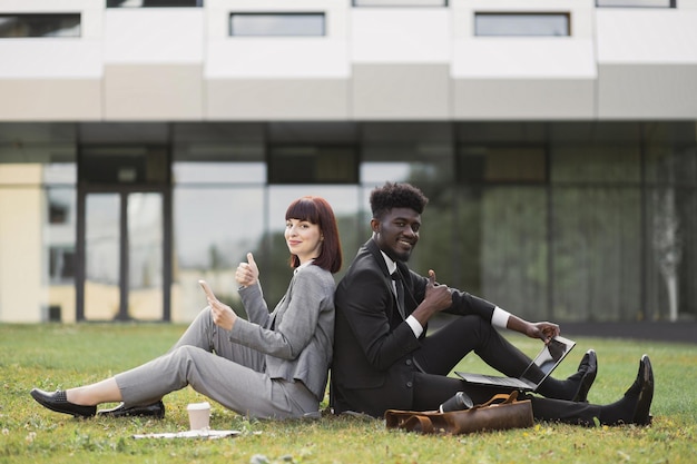 Multiracial male and female partners sitting backtoback on grass near office showing thumb up