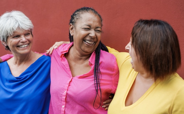 Multiracial mature women hugging each other in the city with red wall in background - Elderly people and friendship concept