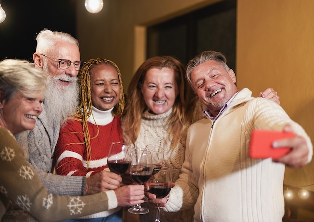 Photo multiracial senior friends cheering with red wine while taking a selfie -