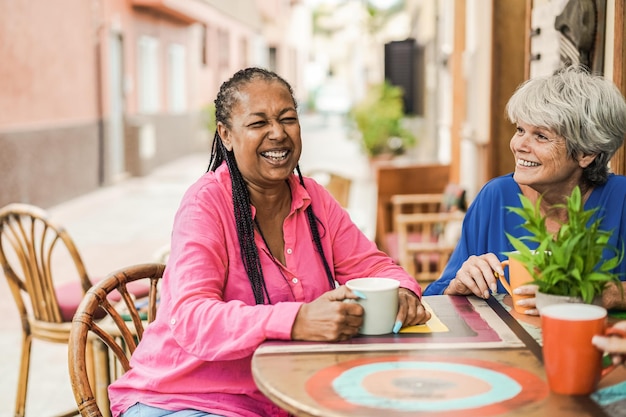 Multiracial senior friends meet and chat at bar outdoor while drinking coffee together - Focus on african woman face