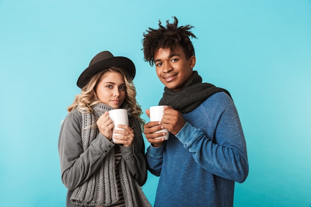 Multiracial teenage couple wearing winter clothes standing isolated over blue wall, holding cups of tea