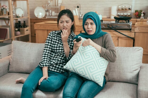 Multiracial young people friendship concept. vintage style
photo of group asian arabic and korean ladies sitting on couch with
scared face. frowning girls watching horror movie on tv at home
kitchen.