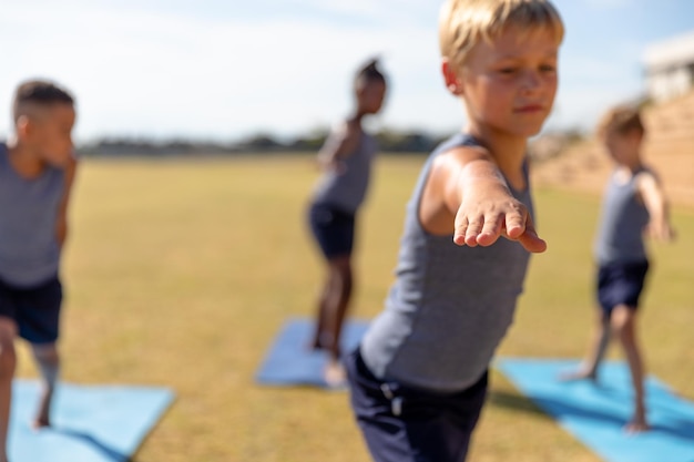 Foto multiraciale basisschooljongens doen krijger 2 pose tijdens het trainen op yogamat tijdens zonnige dag