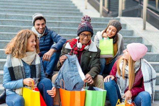 Multiraciale groep vrienden die na het winkelen op de tribune zitten, toont de jongen tevreden zijn aankopen aan de meisjes die een helder kleurenfilter glimlachen