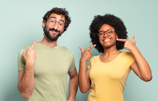 Multiraciale paar vrienden glimlachend vol vertrouwen wijzend op hun eigen brede glimlach, positieve, ontspannen, tevreden houding