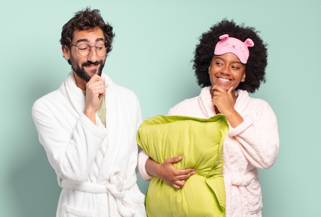 Multiraciale paar vrienden lachend met een gelukkige, zelfverzekerde uitdrukking met de hand op de kin, zich afvragend en opzij kijkend. pyjama's en huisconcept