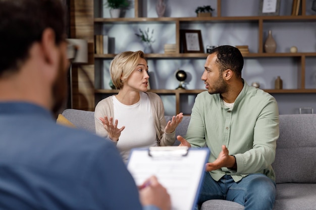 Foto multiraciale ruzie tussen man en vrouw tijdens een sessie met een psychotherapeut een bezoek aan een gezinspsycholoog voor een getrouwd stel een dokter raadpleegt een ongelukkig getrouwd stel help psycholoog