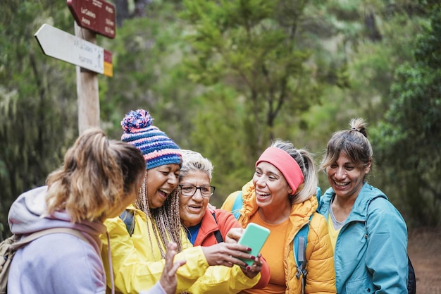 Foto multiraciale vrouwen die plezier hebben met het gebruik van mobiele telefoon voor webkaart terwijl ze het bos in trekken - focus op blank senior gezicht