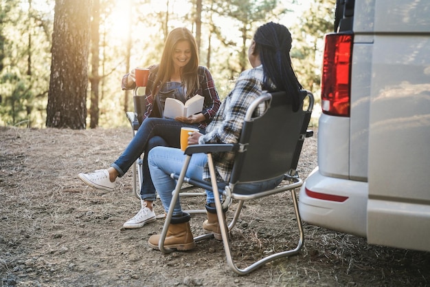 Multiraciale vrouwenvrienden die plezier hebben met kamperen met camper terwijl ze buiten lezen en koffie drinken focus op linker senior hand met boek