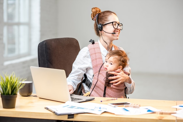Multitasking businesswoman working with headset and laptop sitting with her baby son at the white office interior