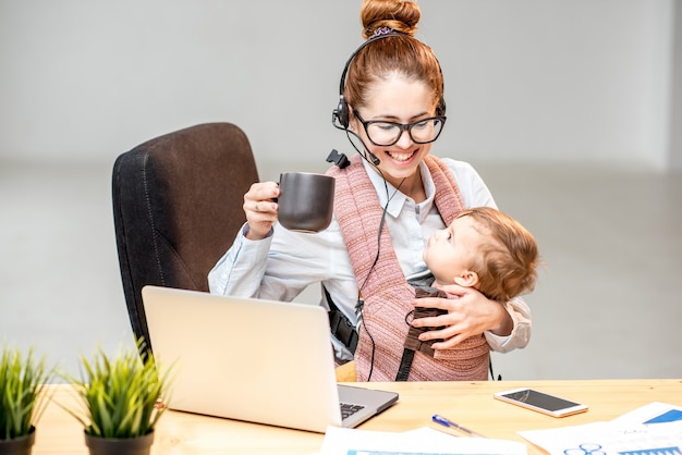 Photo multitasking businesswoman working with headset and laptop sitting with her baby son at the white office interior