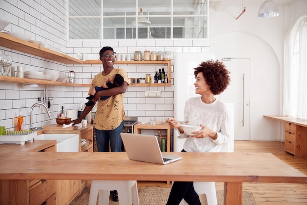 multitasking father holds baby son and cleans surface as mother uses laptop and eats breakfast