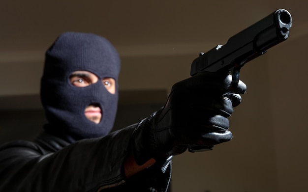 Murderer with balaclava holds gun in leather jacket and gloved hand background