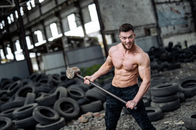 Muscle atleet sterke man shirtless probeert een enorme band te raken met een hamer. Veel banden op de achtergrond in het verlaten magazijn.