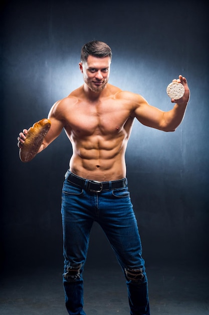 Muscled sportsman holding cereal cracker and bread Choosing between healthy and harmful food naked torso Grey background Closeup