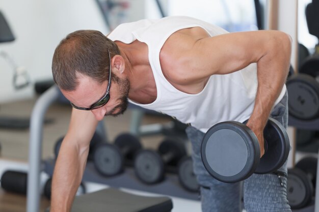 Foto musculaire blanke sterke man die dumbbell rows uitvoert in een fel verlichte sportschool een geconcentreerde man met spieren die werken aan een dumbbell row met één arm hij draagt een witte tank top en zonnebril