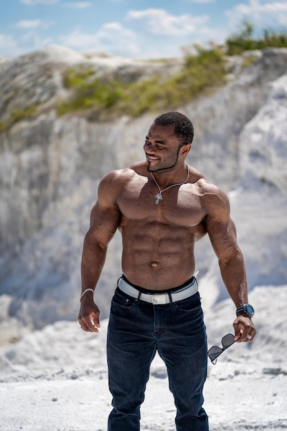 Musculaire zwarte man staat shirtloos op het witte zandstrand met een zonnebril in zijn hand