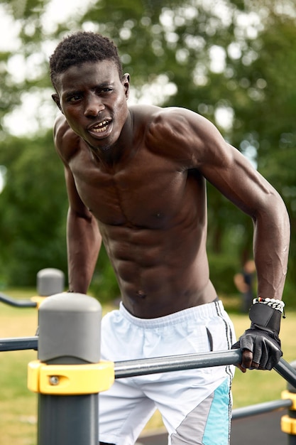 Muscular african american athlete in with a naked sculpted torso doing push-ups on the uneven bars in the urban landscape, Side shot