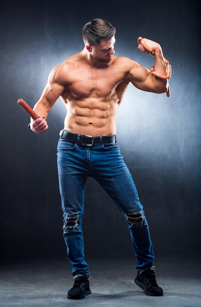 Muscular athlete with naked torso playing muscles with smoked\
sausage in one hand and sausages on a neck full length photo\
lifestyle concept