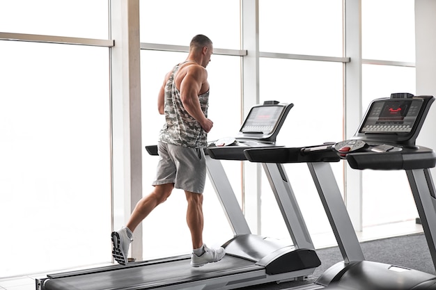 Muscular athletic bodybuilder fitness model running treadmill gym near big window