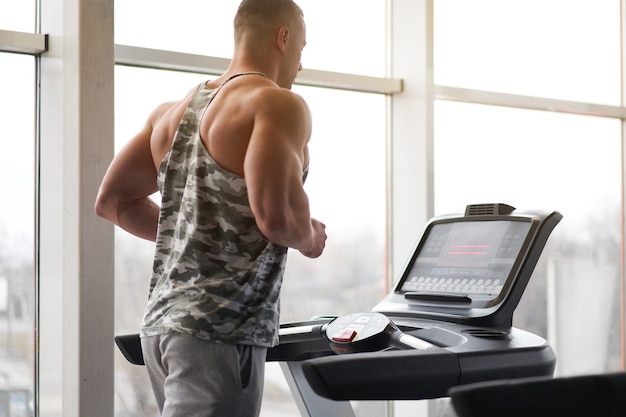 Muscular athletic bodybuilder fitness model running treadmill gym near big window