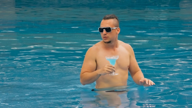 The muscular athletic man drinking blue cocktail alcohol liquor\
in swimming pool at hotel. cocktail bar in the swimming pool.\
portrait of sexy boy in pool outdoor. men with sunglasses.