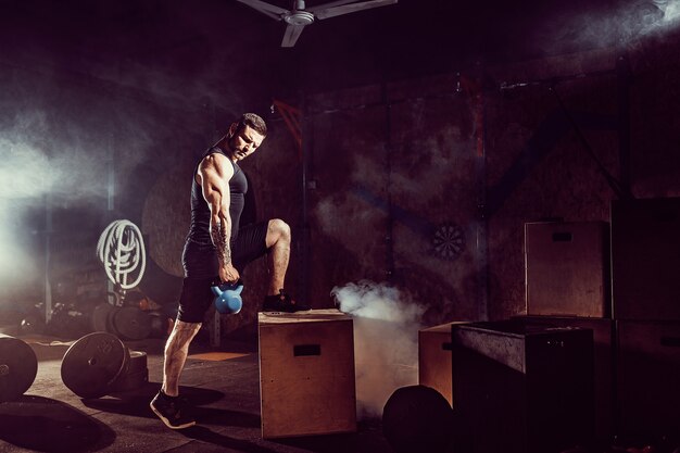 Photo muscular attractive caucasian bearded man lifting two kettlebells in a gym. weight plates, dumbbell and tires in background.