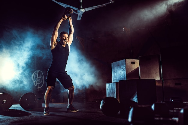 Muscular attractive caucasian bearded man lifting two kettlebells in a gym. Weight plates, dumbbell and tires in background.