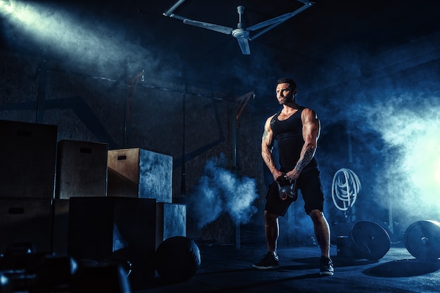 Muscular attractive caucasian bearded tasttoed man lifting kettlebell in a gym. Weight plates, dumbbell and tires in smoke