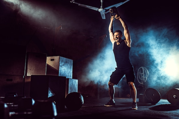 Muscular attractive caucasian bearded tattooed man lifting kettlebell in a gym.