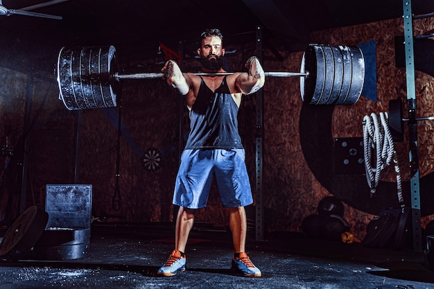 Muscular bearded tattooed fitness man doing deadlift a barbell over his head in modern fitness center. 