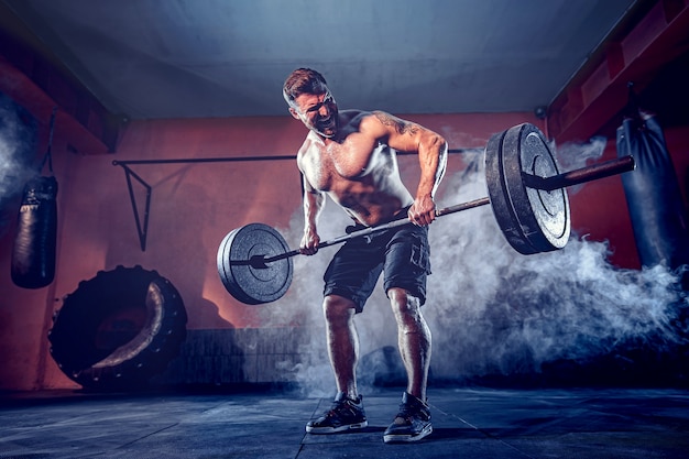 Uomo barbuto muscolare allenandovi in palestra facendo esercizi, torso nudo maschile forte abs. fumo sul muro