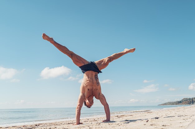 muscular bodybuilder fitness man with a naked torso stands on arm on the beach