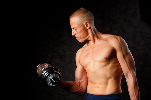 Muscular bodybuilder guy doing exercises with dumbbell over dark background.