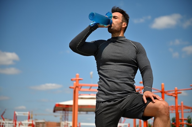 Muscular build handsome middle aged sportsman drinking water while resting after workout on the background of athletic field