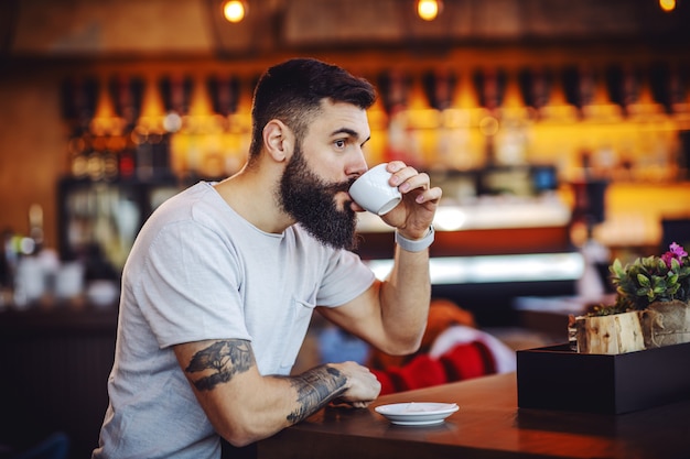 muscular caucasian bearded tattooed hipster sitting in cafe and enjoying his fresh morning coffee.