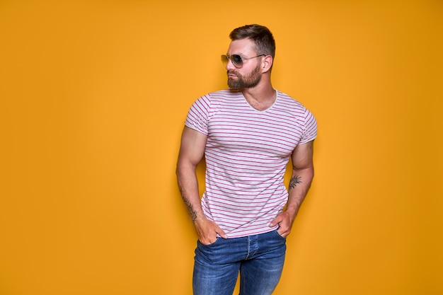 Muscular confident bearded male in shirt jeans and stylish sunglasses standing in studio over yellow.